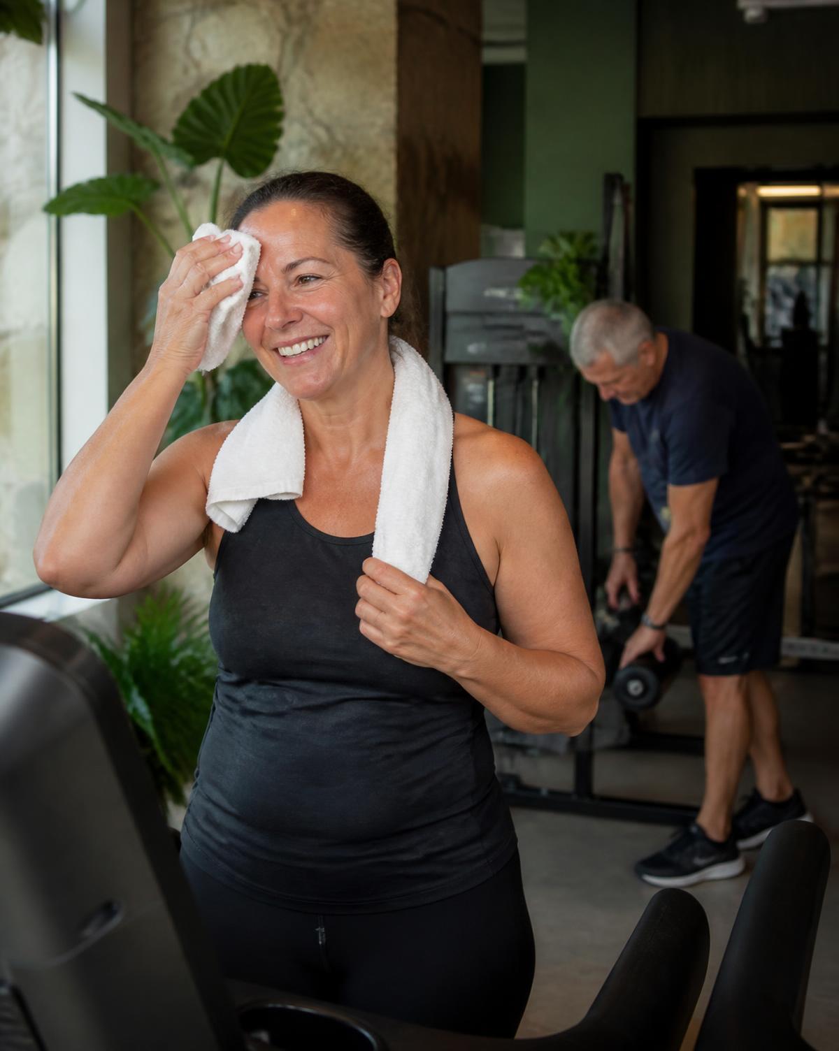 A member in her fifties smiling after a cardio session, with another member training behind her
